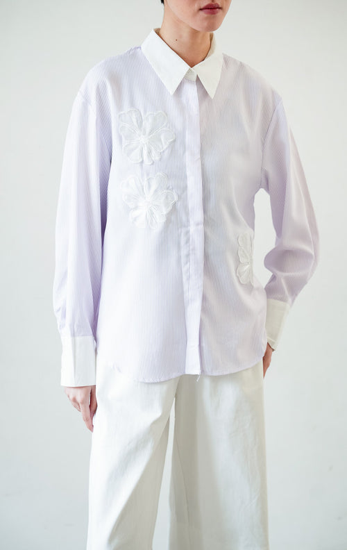 Kemeja wanita lengan panjang motif garis-garis ungu (purple vertical stripes) Wilfred Top dengan detail aplikasi bordir bunga (embroidered floral motifs), kerah dan manset kontras putih (contrasting pointed collar and cuffs), serta bukaan kancing depan. 