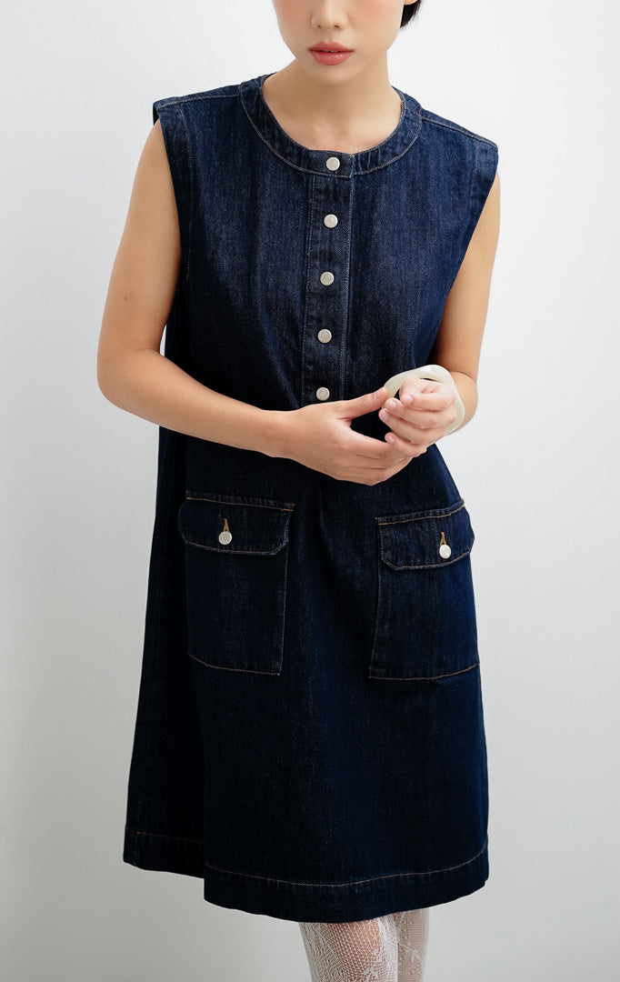 Mini dress wanita tanpa lengan (sleeveless) berbahan denim gelap (dark denim) Winnie Dress dengan kancing jepret (snap-button), saku flap depan, dan saku samping.