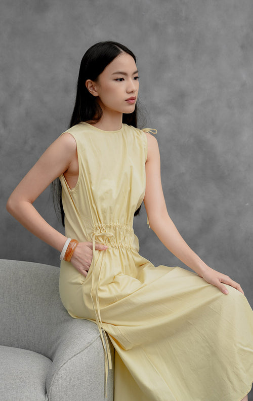 Gaun katun tanpa lengan (sleeveless dress) wanita warna kuning muda (yellow) Winnifred Dress dengan potongan A-line, detail kerut (gathered detailing), aksen tali serut (drawstring), dan bukaan resleting belakang.