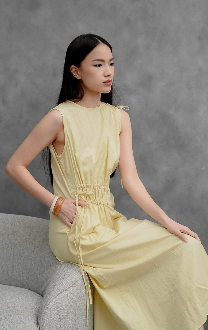 Gaun katun tanpa lengan (sleeveless dress) wanita warna kuning muda (yellow) Winnifred Dress dengan potongan A-line, detail kerut (gathered detailing), aksen tali serut (drawstring), dan bukaan resleting belakang.