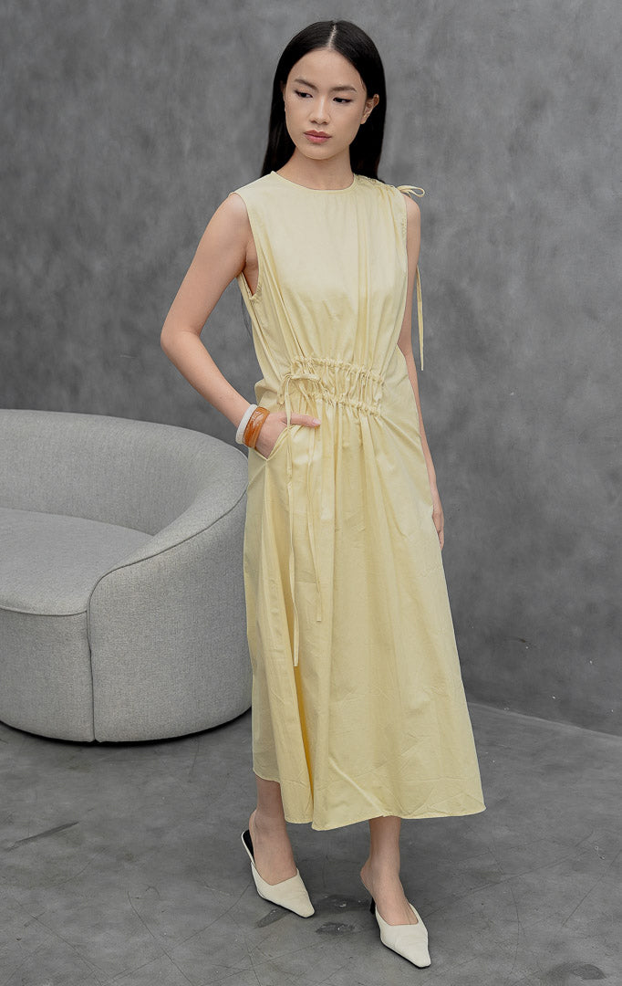 Gaun katun tanpa lengan (sleeveless dress) wanita warna kuning muda (yellow) Winnifred Dress dengan potongan A-line, detail kerut (gathered detailing), aksen tali serut (drawstring), dan bukaan resleting belakang.
