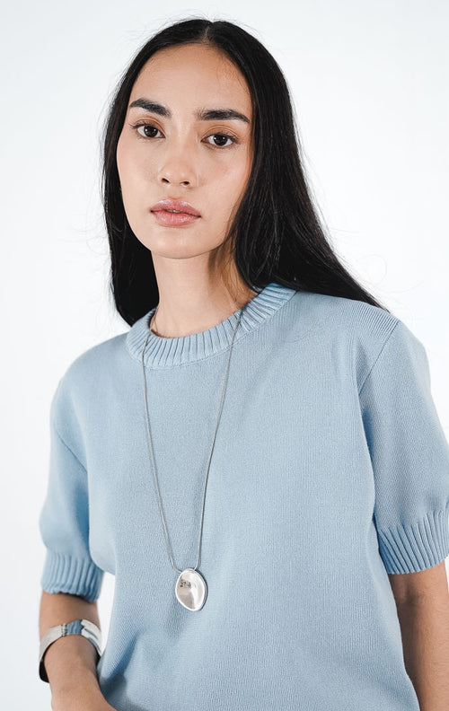 Zaelia Top warna dusty blue, atasan rajut lengan pendek berkerah bulat (crewneck) dengan detail rib, dipadukan dengan kalung liontin perak.