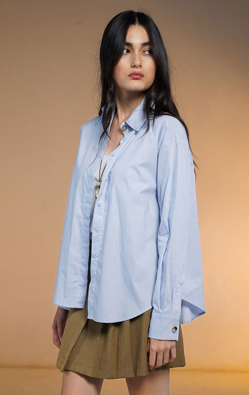 Model wanita memakai Zorrel Top Blue, kemeja lengan panjang warna biru muda dengan kancing statement emas di kerah, dipadukan dengan rok lipit coklat