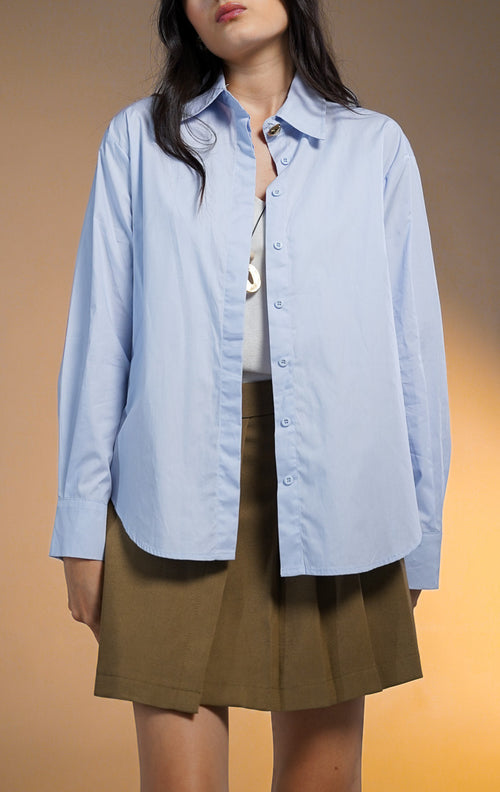 Model wanita memakai Zorrel Top Blue, kemeja lengan panjang warna biru muda dengan kancing statement emas di kerah, dipadukan dengan rok lipit coklat