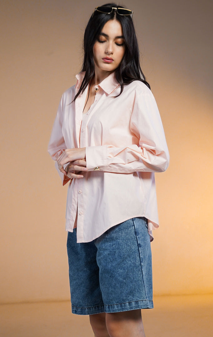 Model wanita memakai Zorrel Top Peach, kemeja lengan panjang warna pink pastel dengan kancing statement emas di kerah, dipadukan dengan celana pendek denim.